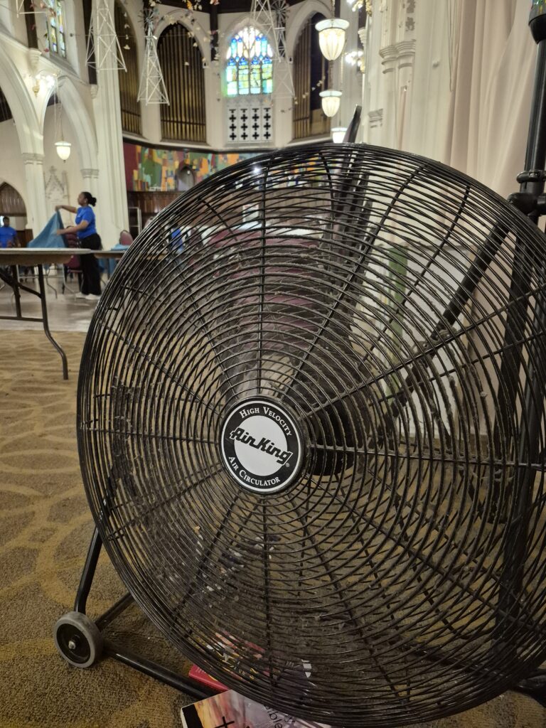 A large fan in the sanctuary dining room with volunteers and stained glass in the background
