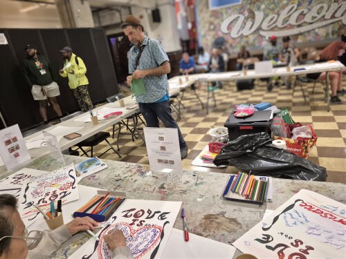 A teaching artist stands in the center of the multipurpose room surrounded by guests at tables completing artwork with the theme Love Is...