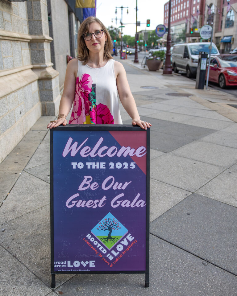 A woman in a white floral dress stands behind a two-tone sign welcoming guests to the Be Our Guest Gala